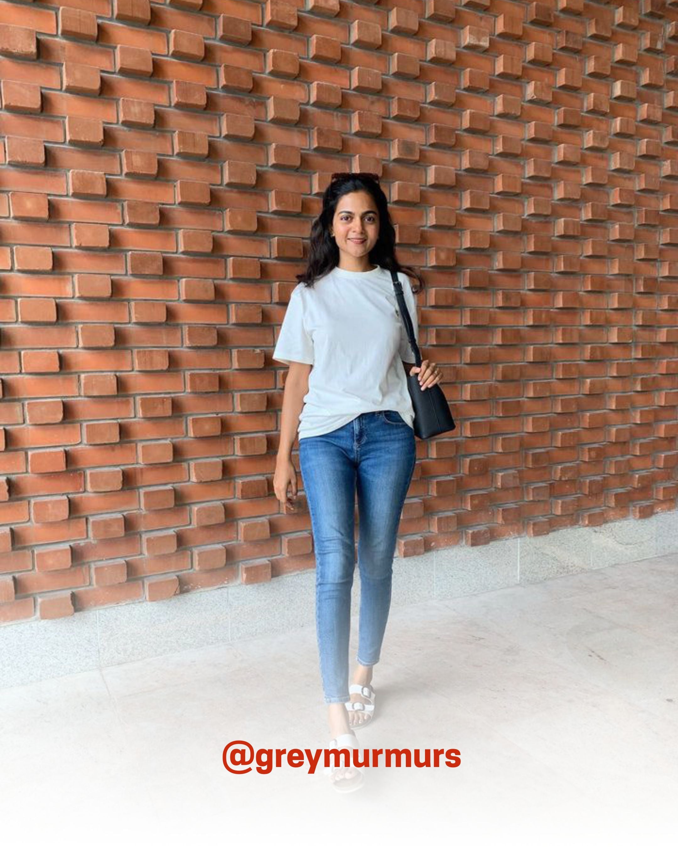 Young woman wearing a white organic cotton T-Shirt with blue jeans, smiling against a brick wall, showcasing 2512’s sustainable t-shirts and eco friendly fashion.